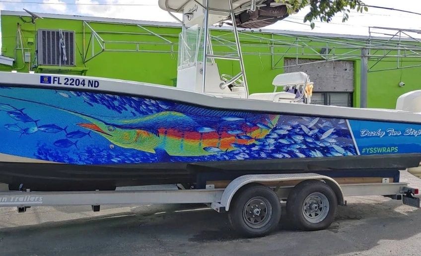 A center-console boat on a trailer showcasing a vibrant custom vinyl hull wrap featuring a colorful sport fish graphic against a deep blue aquatic background.