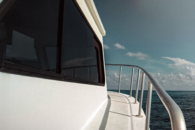 A view along the white exterior deck of a yacht, featuring dark tinted windows and a metal railing looking out over the blue ocean and horizon.