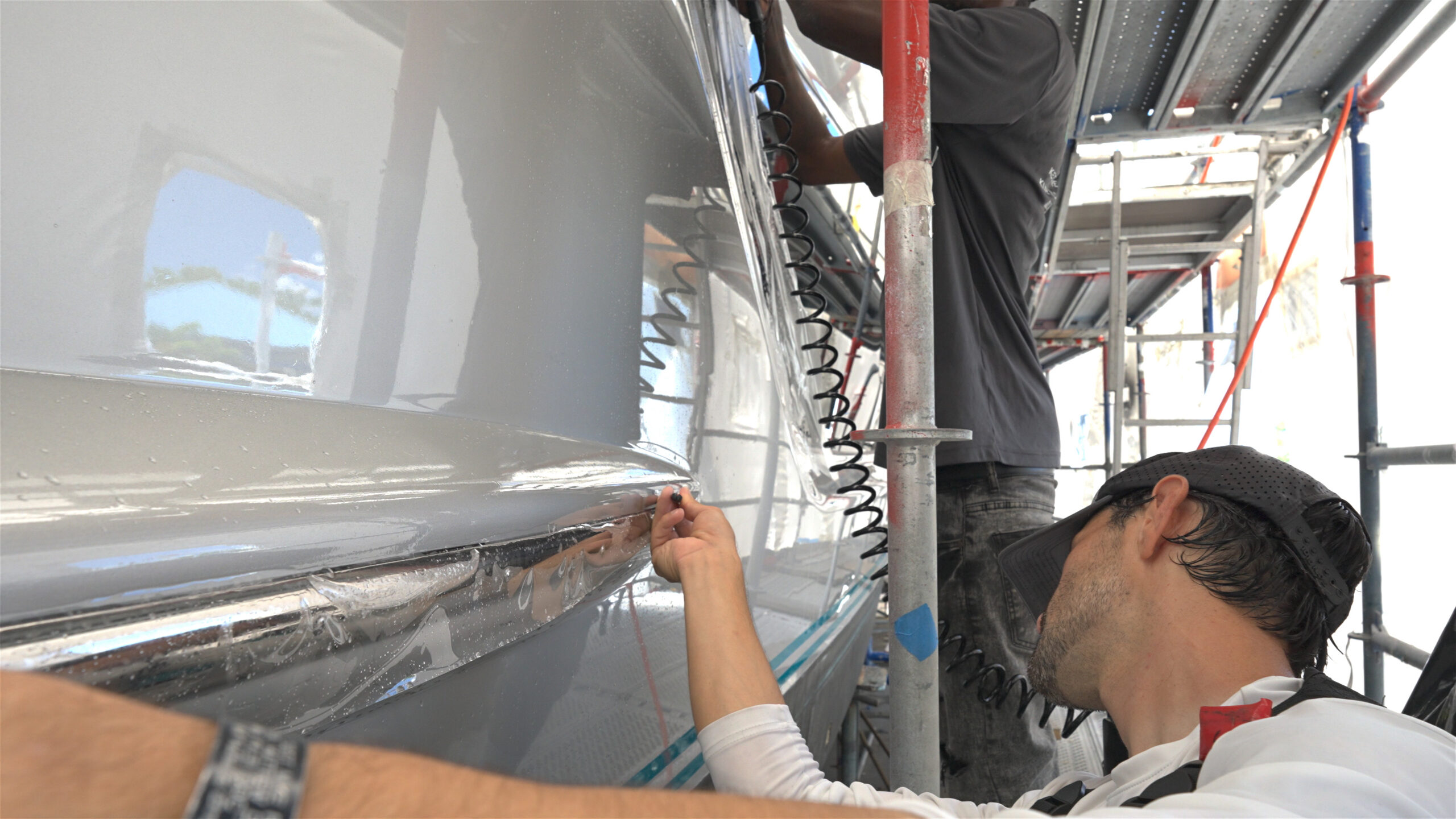 A professional technician carefully applying clear marine paint protection film to the metallic trim and grey hull of a yacht, with a second worker on scaffolding in the background.
