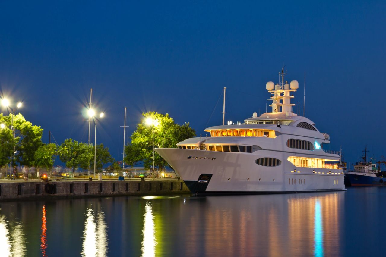 A large, illuminated luxury white superyacht docked at a marina at night, with warm lights reflecting beautifully on the calm water against a deep blue evening sky.